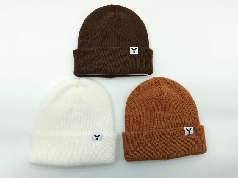 Drei gefaltete Rippenstrick-Beanies mit Umschlagbund in Dunkelbraun, Cremeweiß und Karamellorange, jede mit kleinem aufgenähtem Markenpatch