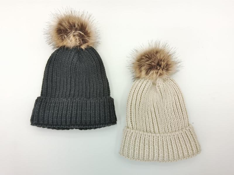 Zwei voluminöse Strickmützen mit großer Kunstfellbommel in Dunkelgrau und hellem Beige auf weißem Hintergrund