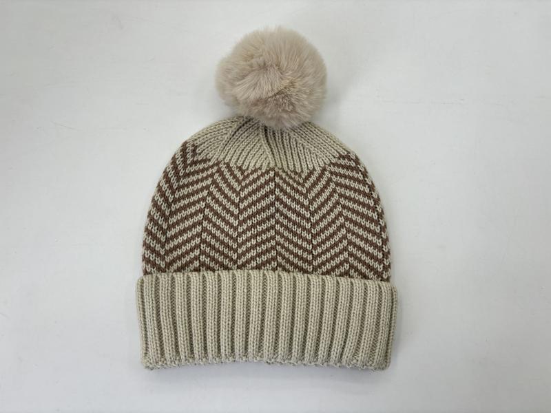 Beige-braune Wintermütze mit Chevron-Strickmuster und großem flauschigem Pompom aus Kunstfell vor weißem Hintergrund