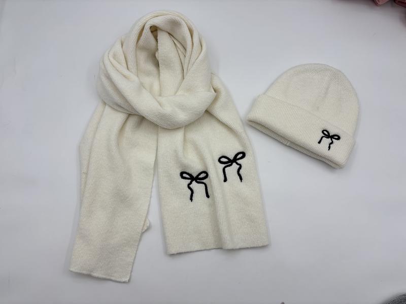 Cremeweißes Rippenstrick-Winterset aus Mütze und Schal mit bestickten schwarzen Schleifenmotiven, flach auf hellem Untergrund