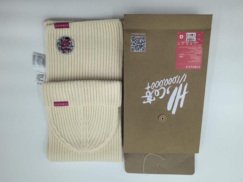 Cremefarbene Rippenstrickmütze und passender Schal mit burgunderrotem Logoaufnäher, präsentiert neben einer Kraftpapier-Geschenkbox