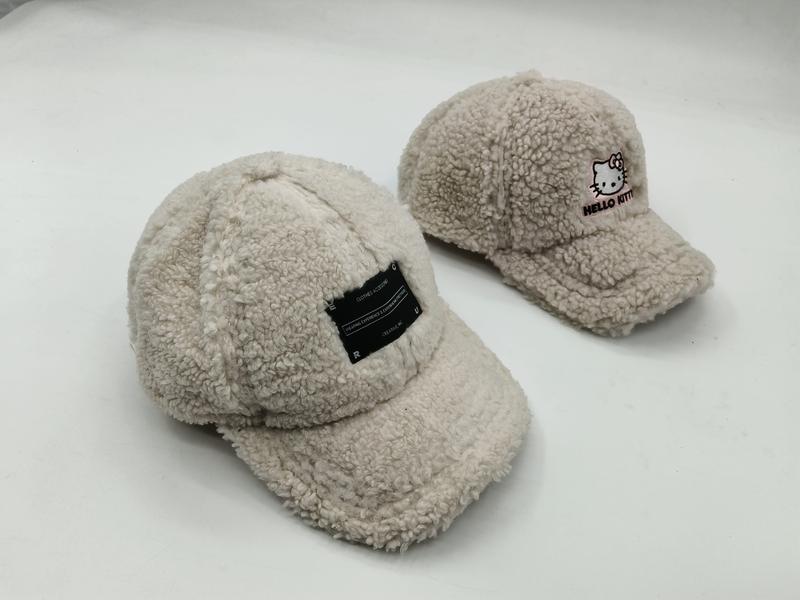 Zwei cremefarbene Teddy-Fleece-Schirmmützen nebeneinander, eine mit schwarzem Label-Aufnäher, die andere mit Hello-Kitty-Stickerei
