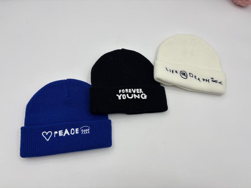 Drei gerippte Umschlagmützen in Blau, Schwarz und Creme mit bestickten englischsprachigen Slogans Peace, Forever Young und Life or Death