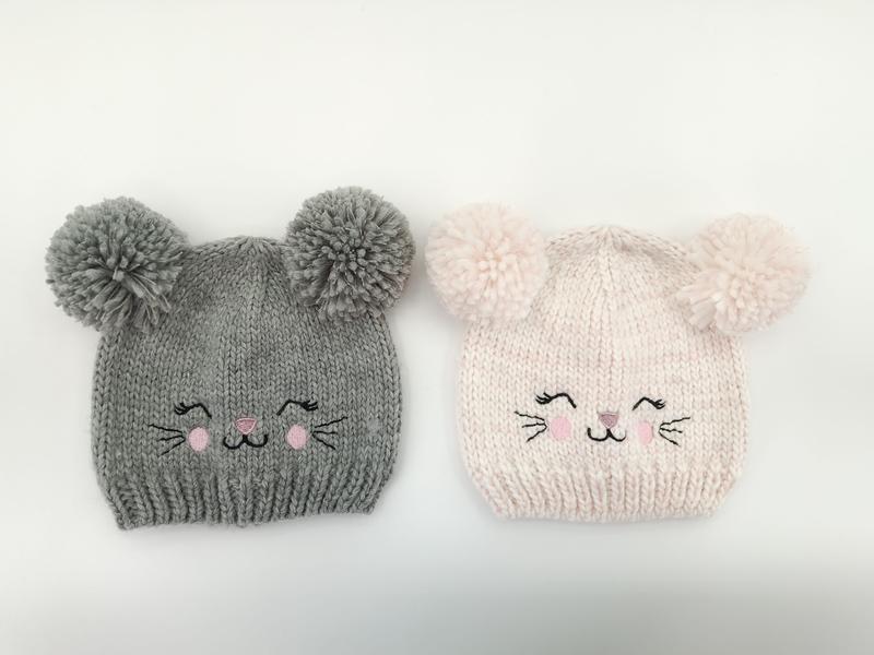 Zwei handgestrickte Babymützen mit flauschigen Bommel-Ohren und aufgestickten Katzengesichtern, eine in Grau und eine in Hellrosa, auf weißem Hintergrund