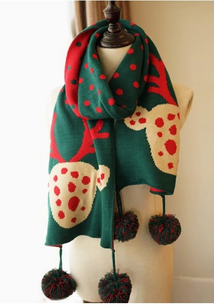 Grüner Strickschal mit Rentier-Allover-Muster und roten Punkten auf einer Schaufensterpuppe, mit dekorativen Pompom-Fransen an den Enden