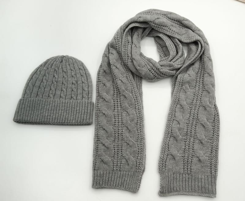 Passendes Winterset aus grauer Zopfstrickmütze und langem Schal im gleichen Flechtmuster, nebeneinander auf weißem Hintergrund
