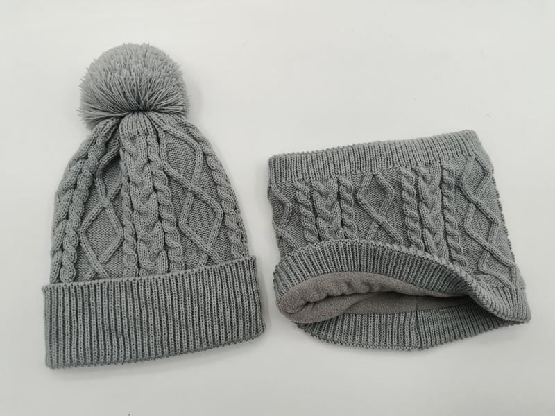 Graue Zopfstrick-Wintermütze mit Pompon und dazu passender Fleece-gefütterter Schlauchschal, freigestellt auf weißem Hintergrund