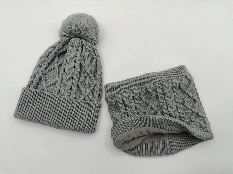 Graue Zopfstrick-Wintermütze mit Bommel und passender Fleece-gefütterter Schlauchschal auf weißem Hintergrund