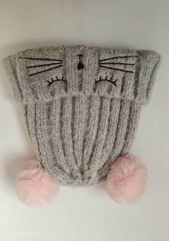Graue gestrickte Wintermütze mit aufgesticktem Katzengesicht, Schnurrhaaren und zwei pinken Kunstfell-Pompons an der Krempe