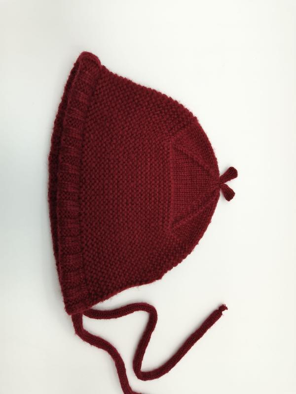 Handgestrickte Babymütze in kräftigem Dunkelrot mit Perlmuster-Textur, Rüschenkante, Kinnbändern zum Binden und charakteristischer Spitze, auf weißem Hintergrund liegend