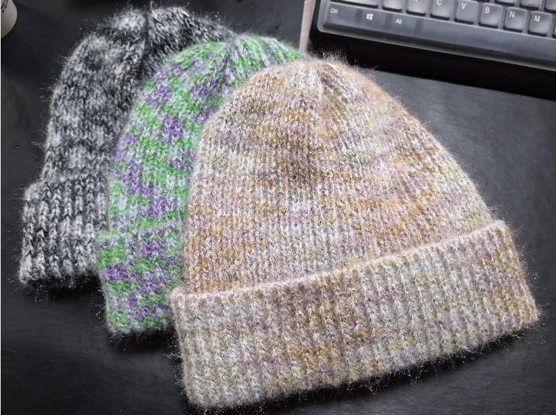 Drei unterschiedliche handgestrickte Wintermützen in Graumeliert, Grün-Lila-Streifen und warmen Beige-Gold-Tönen auf dunklem Untergrund