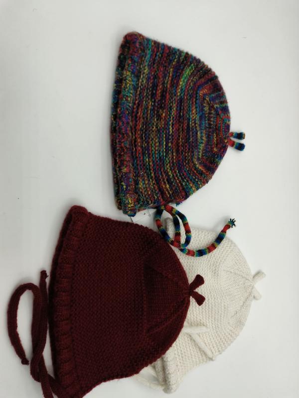 Drei handgestrickte Babyhäubchen mit Kinnbändern nebeneinander in buntem Farbverlaufsgarn, satten Burgunderrot und klassischem Cremeweiß, auf weißem Untergrund