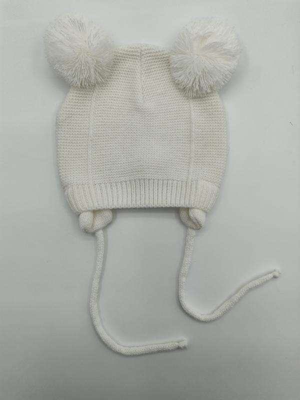 Weiße gestrickte Babymütze mit zwei flauschigen Bommeln oben und langen Kinnbändern, flach auf weißem Untergrund fotografiert