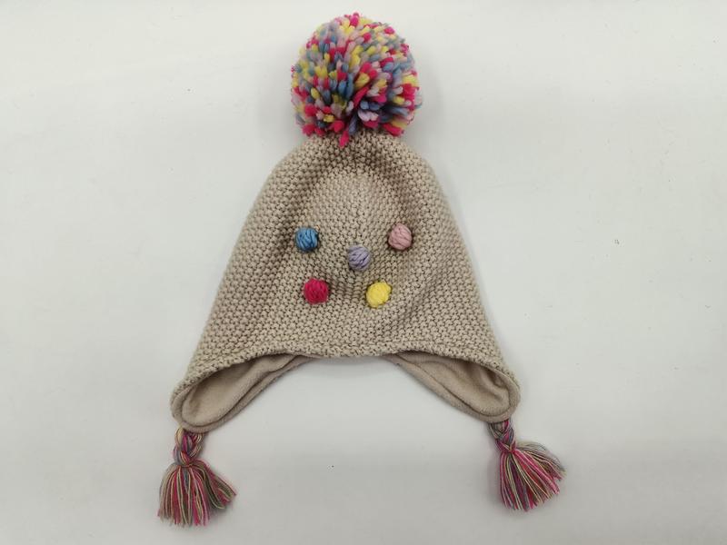 Beige gestrickte Kindermütze mit Ohrenklappen, großem buntem Bommel oben, farbigen Knopfakzenten und geflochtenen Zierquasten