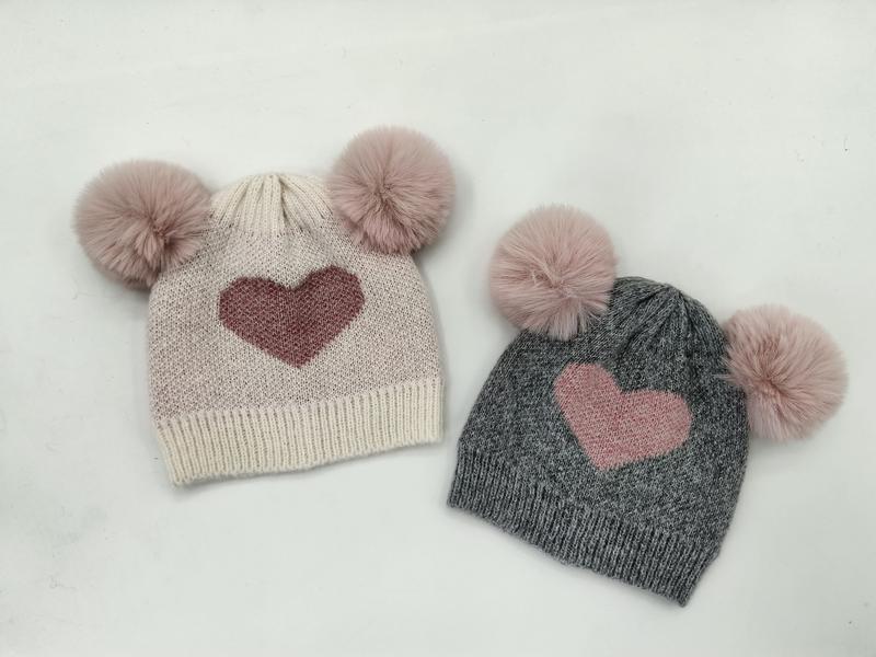 Flauschige Kindermützen aus Strickstoff in Creme und Grau mit zwei Kunstfell-Pompons und eingestricktem Herzmotiv, nebeneinander auf weißem Hintergrund