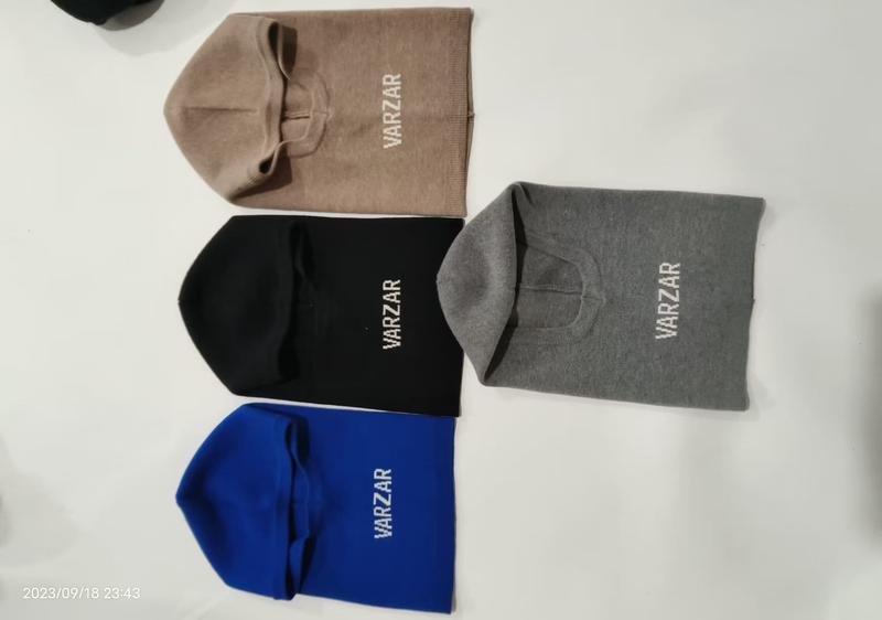 Vier gestrickte Sturmhauben in Beige, Schwarz, Grau und Blau flach auf weißer Fläche, mit besticktem Markenlogo