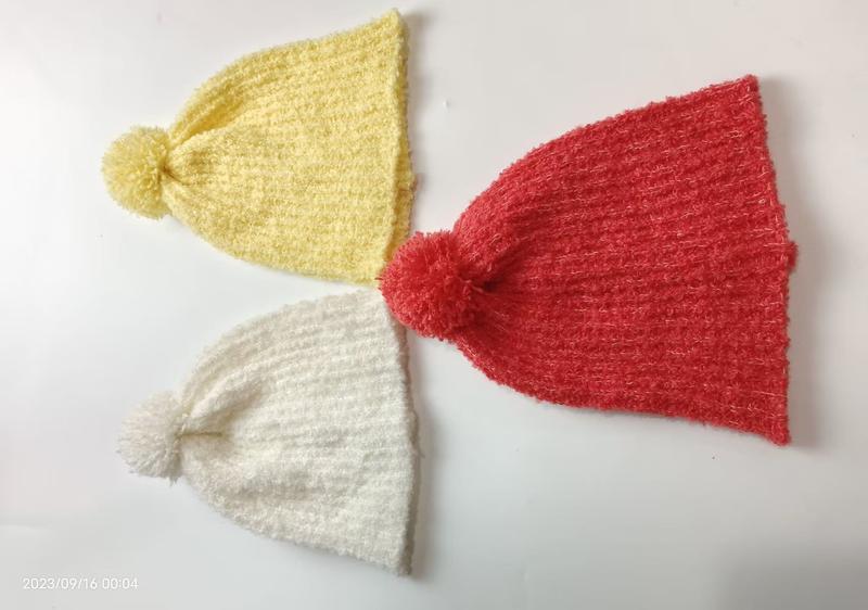 Drei gestrickte Bommelmützen in Sonnengelb, Korallenrot und Reinweiß nebeneinander liegend, flauschige Wintermützen im Flatlay