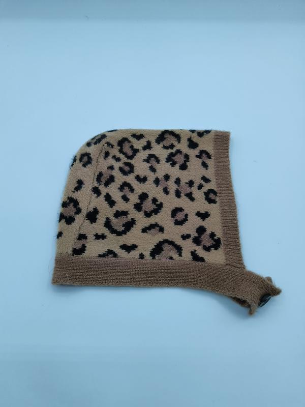 Gestrickte Säuglingsbonnetmütze im Leopardenprint mit beigem Hintergrund und schwarzen Flecken, mit Rippenrand und Kinnbändchen – flach auf weißem Untergrund