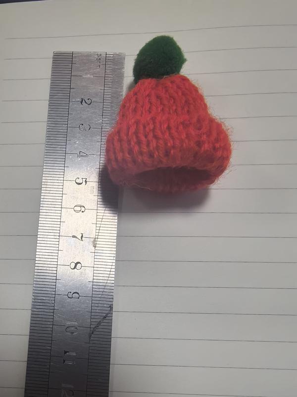 Winzige handgestrickte Wollmütze in Rot mit grünem Bommelpunkt, neben einem Lineal mit ca. 4 cm Höhe