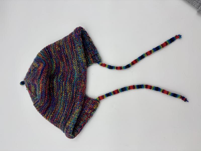 Handgestrickte Babyhäubchen aus mehrfarbigem Buntgarn mit Ohrenschutz und geflochtenen Bindebändern, Flachlage auf weißem Grund
