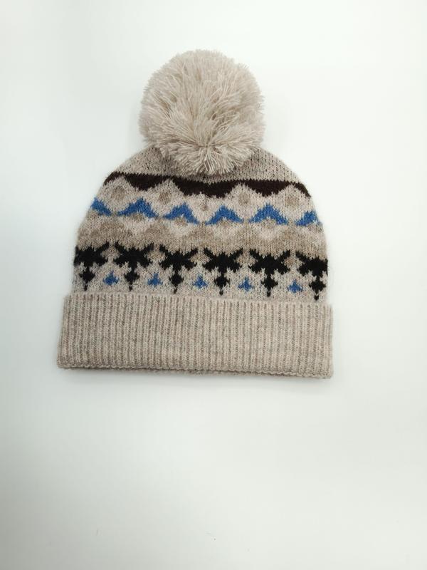 Haferfarben gestrickte Mütze mit blau-schwarzem Schneeflocken-Jacquardmuster und großem cremefarbenem Bommel auf der Kuppe