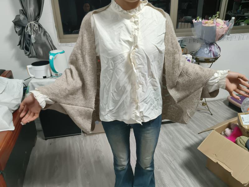 Frau trägt übergroßen beigen Strickjacke über cremefarbenem wattiertem Rüschenkragenhemd mit blauen Schlaghosen in Innenraum