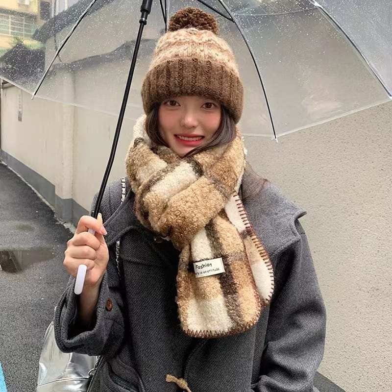 Damen-Winterset bestehend aus flauschigem Sherpa-Schal im Karomuster und grob gestrickter Pudelmütze in Braun-Creme unter Regenschirm
