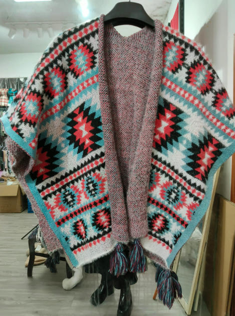 Offener Strickponcho mit Aztekenmuster in Rot, Petrol, Schwarz und Weiß sowie bunten Quastenfransenabschlüssen auf Kleiderbügel