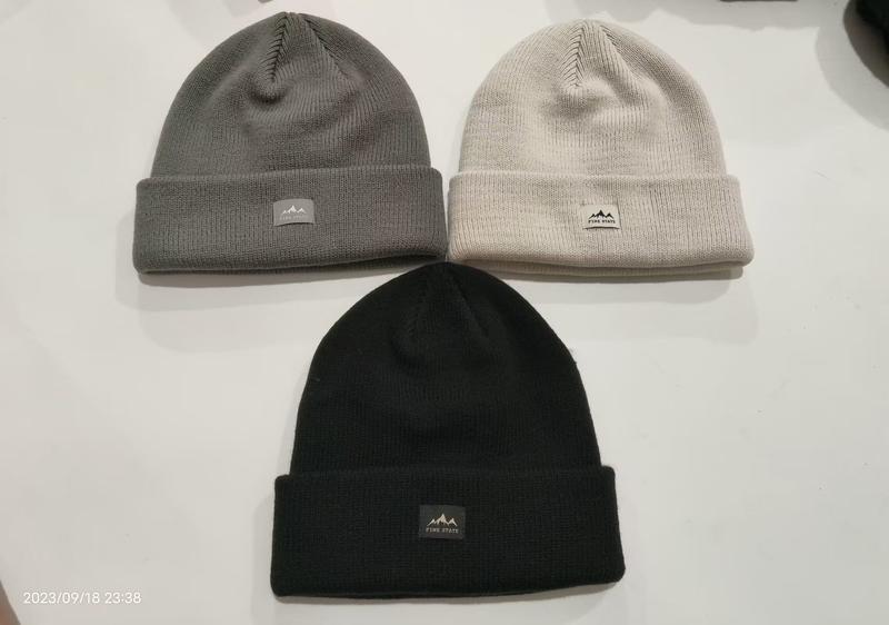 Drei gerippte Strickmützen mit Umschlagbund in Schwarz, Grau und Cremeweiß flach liegend, mit Berglogo-Patch