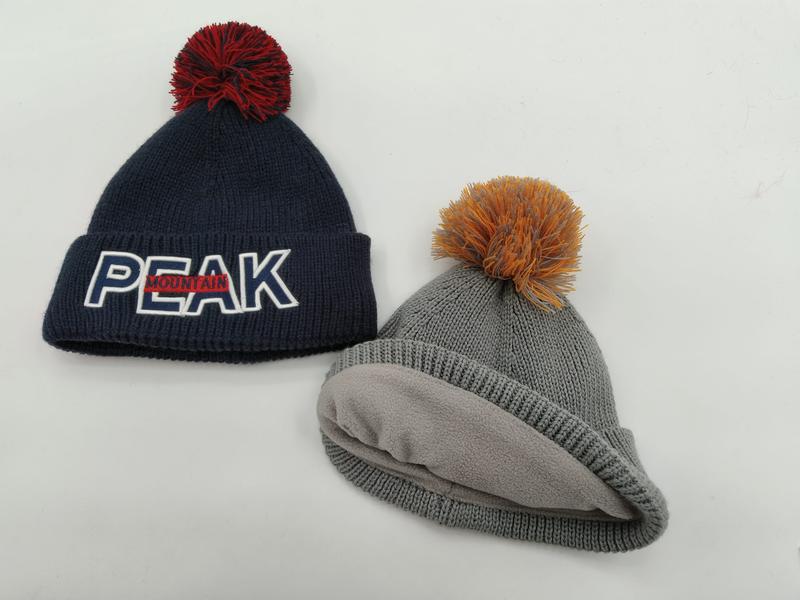 Zwei Strickmützen mit Bommeln: Navy-Blau mit Peak Mountain Logo und rotem Bommel, graue Fleece-gefütterte Mütze mit orangem Bommel, auf weißem Hintergrund