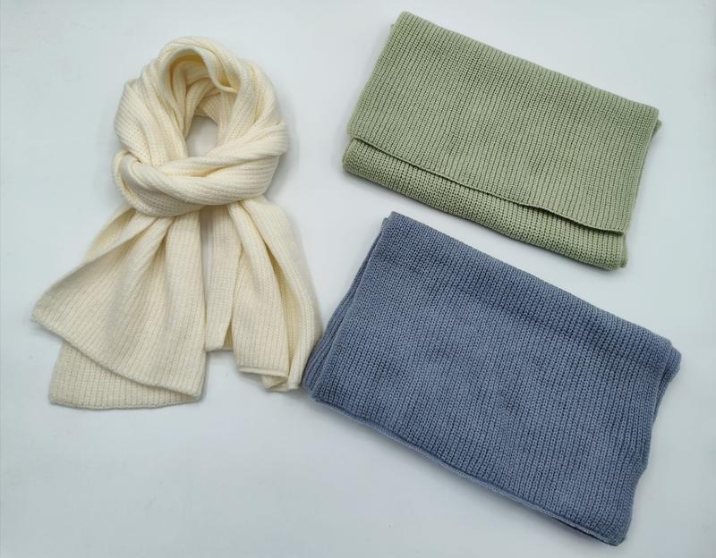 Drei winterliche Strickaccessoires in Creme, Salbeigrün und Stahlblau: locker gebundener langer Schal und zwei zusammengelegte Schlaufsnoods auf hellem Untergrund