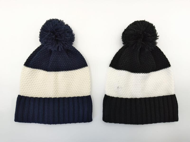 Zwei Rippenstrick-Pompom-Mützen nebeneinander – links Marine-Blau mit Cremstreifen, rechts Schwarz mit Cremstreifen – auf weißem Hintergrund