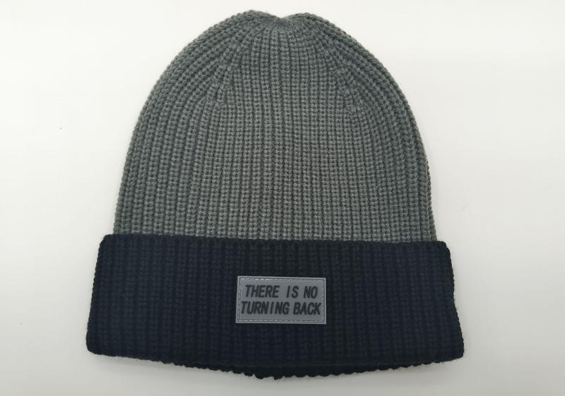 Zweifarbige Wintermütze in Grau und Schwarz mit grobem Rippenmuster und Gummi-Labelaufnäher mit Schriftzug auf dem dunklen Umschlag