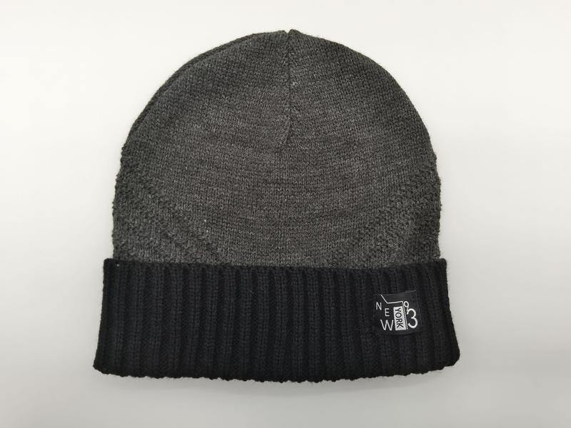 Grau gestrickte Beanie mit kontrastreichem schwarzem Rippenumschlagbund und eingenähtem Schriftzug-Label auf weißem Hintergrund