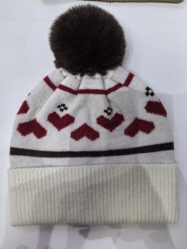 Weiße Strickmütze mit roten Herzchen-Muster, dunkelbraunem Kunstfell-Pompon und geripptem Umschlagrand auf weißem Hintergrund