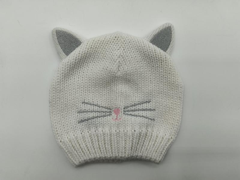 Weiße grob gestrickte Baby-Beanie mit silber glitzernden Katzenohren, aufgestickten Schnurrhaaren und rosa Nase auf grauem Hintergrund