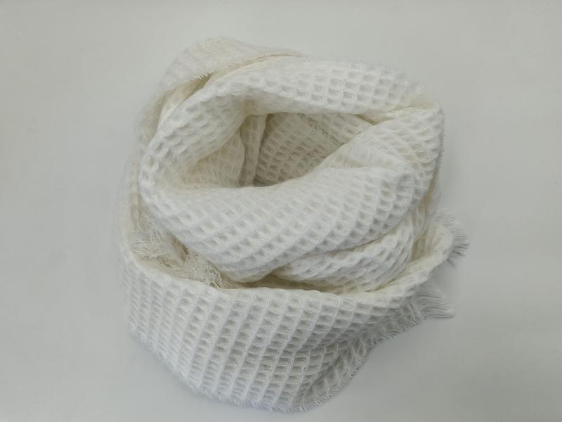 Weißer Schal in Waffeloptik aufgerollt auf heller Fläche mit sichtbaren Fransen und offenem Strickmuster